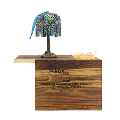 A miniature lamp ornament with a stained glass shade featuring the wisteria pattern of blue, yellow, and purple hues on a bronze colored base with a blue ribbon on top of the lampshade. The lamp ornament is on top of a wooden crate packaging with the engraved text 