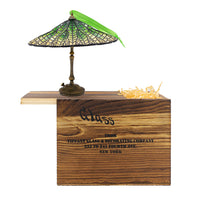 A miniature decorative lamp with a green stained glass shade with a bronze colored base on a wooden crate with the engraved text 