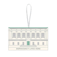 A keepsake ornament featuring the facade of The New York Historical Building. The ornament is mostly a cream white color with silver and green accents and the text 'Democracy Lives Here' at the bottom of the building