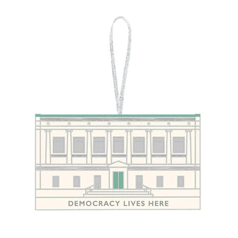 A keepsake ornament featuring the facade of The New York Historical Building. The ornament is mostly a cream white color with silver and green accents and the text 'Democracy Lives Here' at the bottom of the building