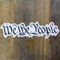 A die-cut sticker featuring the phrase 'We The People' in a black Gothic or Old English style font displayed flat on a rustic brown wooden surface.