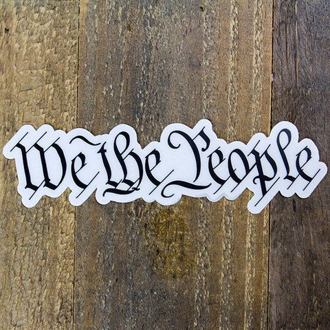 A die-cut sticker featuring the phrase 'We The People' in a black Gothic or Old English style font displayed flat on a rustic brown wooden surface.