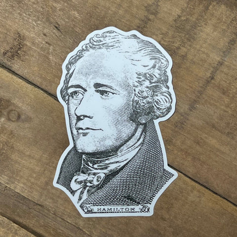 A die-cut sticker featuring a black and white portrait of Alexander Hamilton, placed on a brown wooden surface.