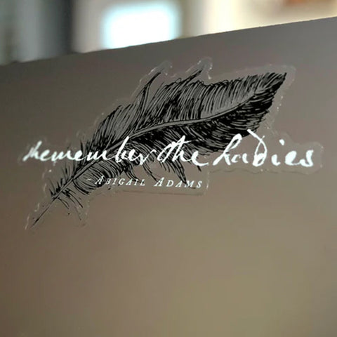 A close-up of a laptop cover featuring a black feather illustration and the title 'Remember the Ladies' in white script, with 'Abigail Adams' printed below.
