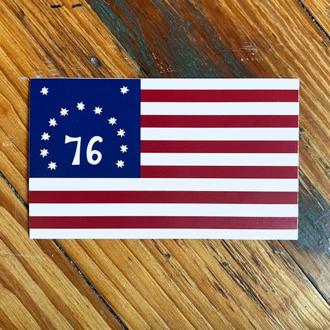 A rectangular Bennington Flag sticker with 13 stars in a circle around the number 76, placed on a wooden surface.