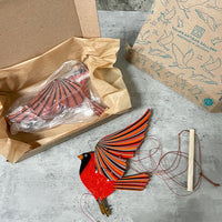 A handmade wooden cardinal bird mobile with red, orange, and black striped wings and body, shown unpackaged next to its box and a second printed box on a gray stone surface.