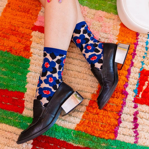 A person's legs wearing black leather block-heel shoes and blue floral socks with a leopard print, sitting on a colorful striped rug.