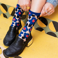 A person wearing patterned crew socks with blue floral motifs over a beige base, paired with black leather loafers and metallic block heels, seated on a yellow and black patterned bench.