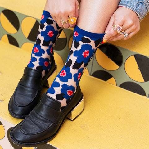 A person wearing patterned crew socks with blue floral motifs over a beige base, paired with black leather loafers and metallic block heels, seated on a yellow and black patterned bench.