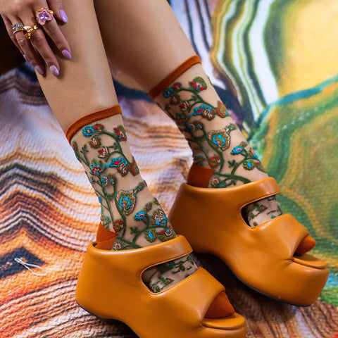 A close-up lifestyle shot of a person's legs wearing sheer beige socks with a green and blue floral vine pattern, paired with bright orange platform slide sandals, resting on a colorful patterned blanket.