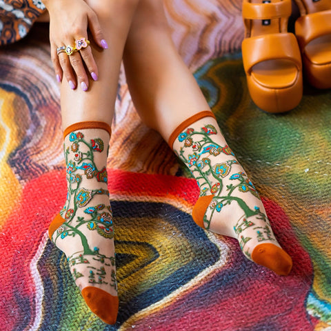 A person wearing patterned ankle socks with orange toes and heels, seated on a colorful rug with tan sandals nearby.