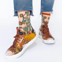 A person wearing low-top sneakers with a glossy brown and gold abstract pattern, white soles, and brown laces, paired with frayed blue jeans and patterned socks.