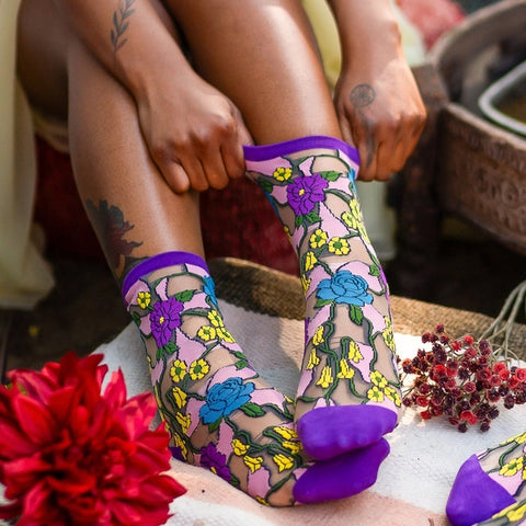 A close-up lifestyle shot of a person sitting and pulling up a pair of sheer patterned socks. The socks feature a light pink background with a colorful floral print in blue, purple, yellow, and green, finished with solid purple cuffs and toes.