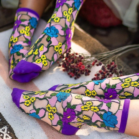 A pair of sheer black knee-high socks with a colorful floral lace-like pattern in pink, blue, yellow, and purple, worn on feet with a decorative background.