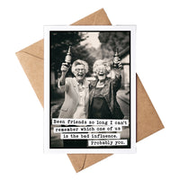 A black-and-white greeting card with a humorous quote at the bottom, shown with a brown kraft envelope on a white background. The card features a photo of two elderly women holding up beer bottles with the text "Been friends so long I can't remember which one of us is the bad influence. Probably You"