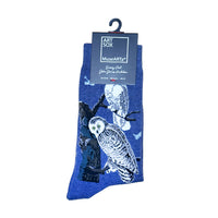 A pair of blue socks with a printed design of two owls perched on tree branches against a night sky, hanging from a black hanger with an Art Sox MuseArts tag.