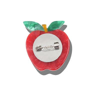 An apple-shaped safety pin brooch with a red body, green leaves, and a yellow stem,  with its silver metal pin back visible.