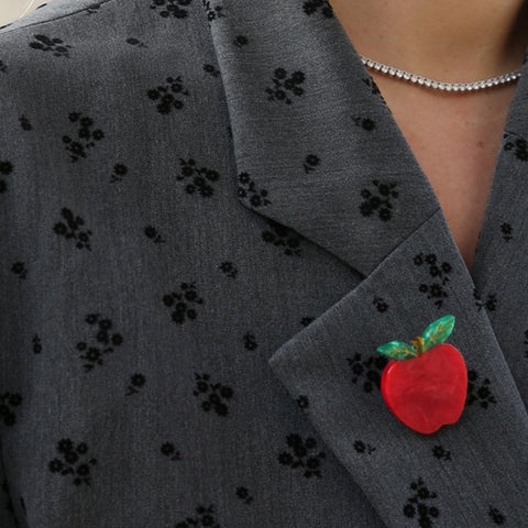 Close-up of a person wearing a gray blazer with a black floral pattern and a red apple brooch on the lapel.