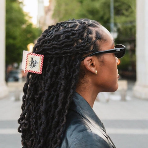 A side-profile portrait of a woman with long black dreadlocks wearing a red and white checkered hair clip with a black graphic, set against a blurred outdoor city background.