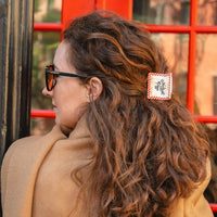 A woman with long, wavy brown hair is shown in profile wearing a small square hair clip. The clip has a white background with a red checkered border and black calligraphic text in the center.