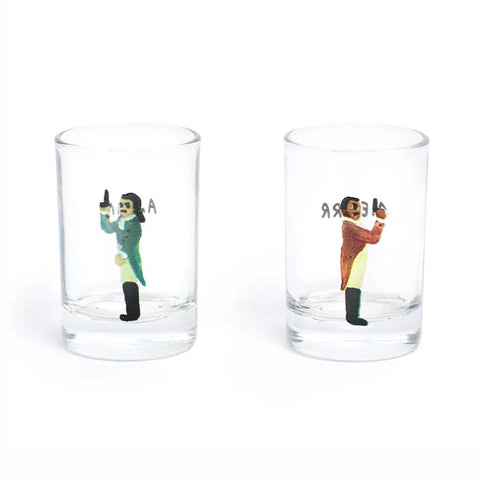 Two clear shot glasses on a white background, each printed with a small illustration of men in time-period clothing
and black text.