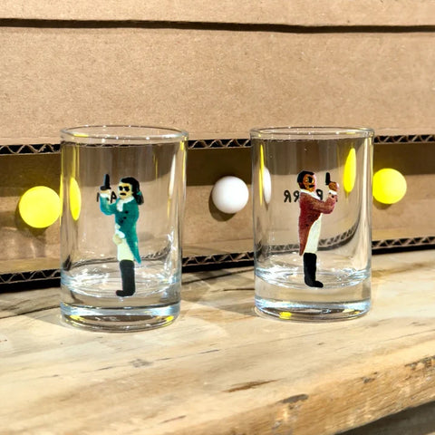 Two clear shot glasses with printed illustrated figures of a man in a teal coat and a man in a red coat, both holding guns, displayed on a wooden shelf in front of a cardboard backdrop with yellow and white balls.