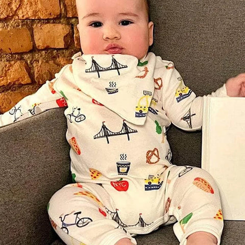 A baby wearing a white-colored long-sleeve footed pajama set with a white triangular bandana bib featuring a colorful New York City-themed print with illustrations of a bicycle, hot dog, pizza, coffee cup, bridge, taxi, and red NYC logos.