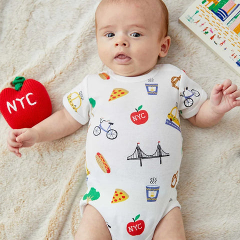 A baby in a white body suit with New York icons as a scattered print in the colors yellow, blue, green, red, blue, and black.