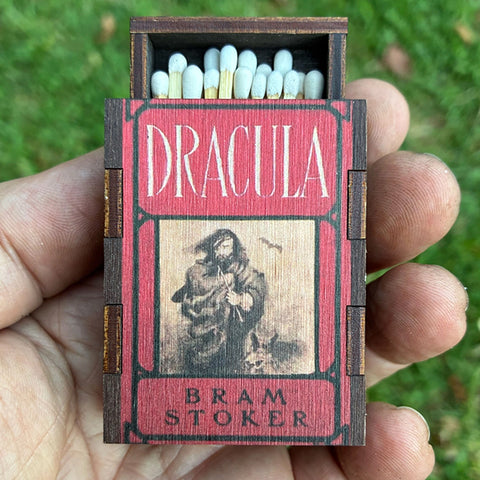 A hand holds a miniature wooden matchbox styled like a Dracula book cover, with match heads visible in the open top. The box is red with white 'DRACULA' text and a sepia-toned illustration, against a blurred green grass background.