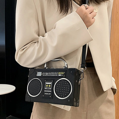 A person wearing a cream blazer and beige pants carries a black rectangular crossbody bag designed to look like a vintage boombox, with silver-tone handles and speaker grilles.