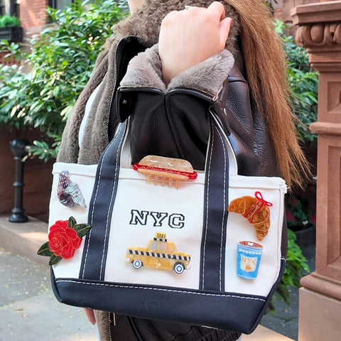 A person wearing a brown faux-fur collar jacket carrying a white canvas tote bag with navy blue trim. The bag features embroidered details, including 'NYC' text, with an array of pins like a yellow taxi, a red rose, a croissant, and a cupcake