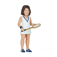 A side view of a woman with brown hair wearing eyeglasses and a white tennis dress with a blue v accent collar holding a tennis racket with blue tennis sneakers and light blue laces 